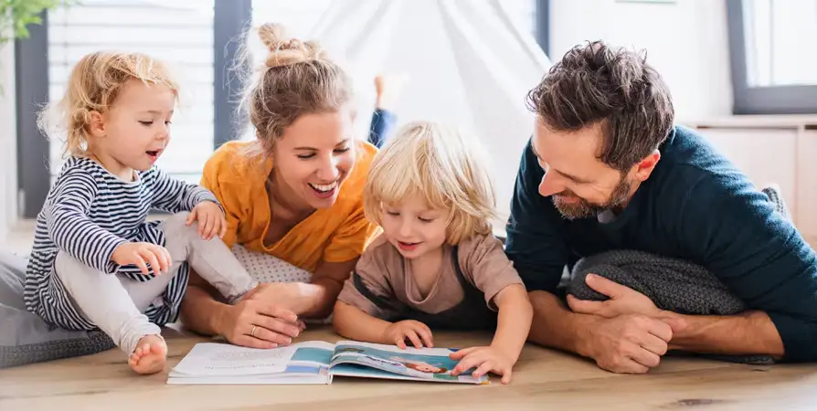 Happy parents with two young children laying on hardwood floors reading a book